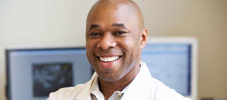 A smiling African American adult male wearing a white collared shirt is seen from the shoulders up. The background is blurred but two computer monitors are visible.